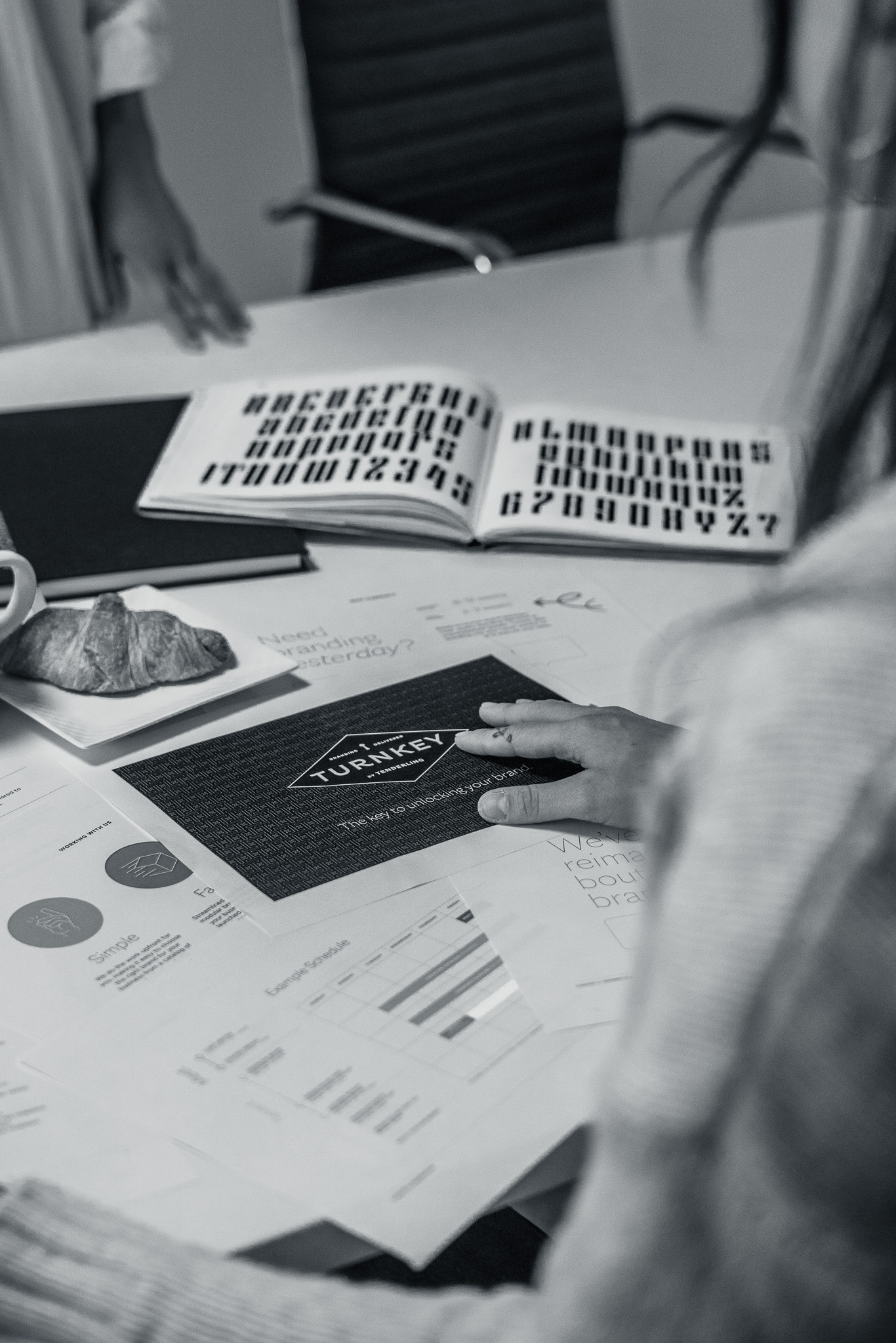 A person reviews business documents and marketing materials at a desk, surrounded by charts, graphs, and an open booklet with various fonts.