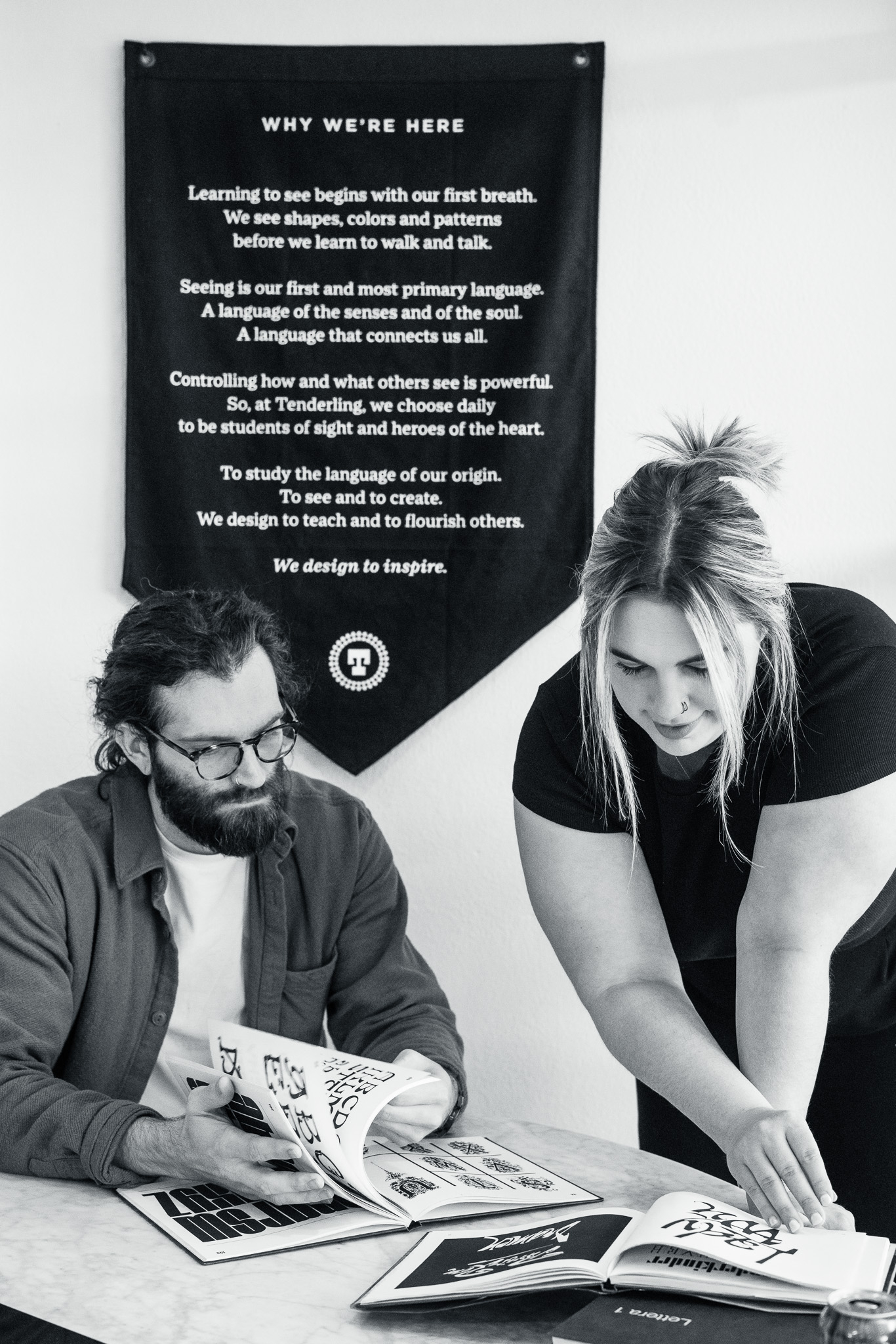 Two people look at open books on a table. A woman points to a page while a man sits beside her. A poster with text about learning and inspiration hangs on the wall behind them.