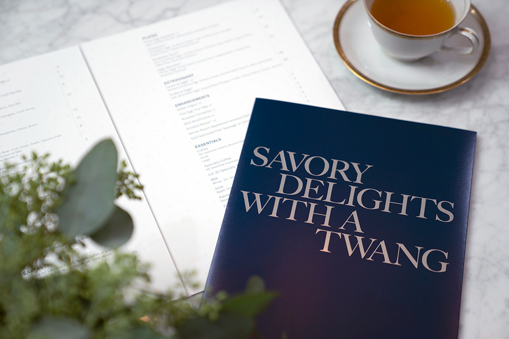 A menu titled "Savory Delights With a Twang" sits on a marble table beside an open menu, a teacup with tea, and some greenery in the foreground.