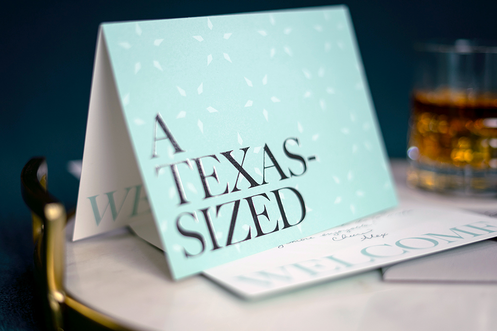 A light blue card with bold black text reading "A TEXAS-SIZED" is displayed on a table next to a glass of brown liquid.
