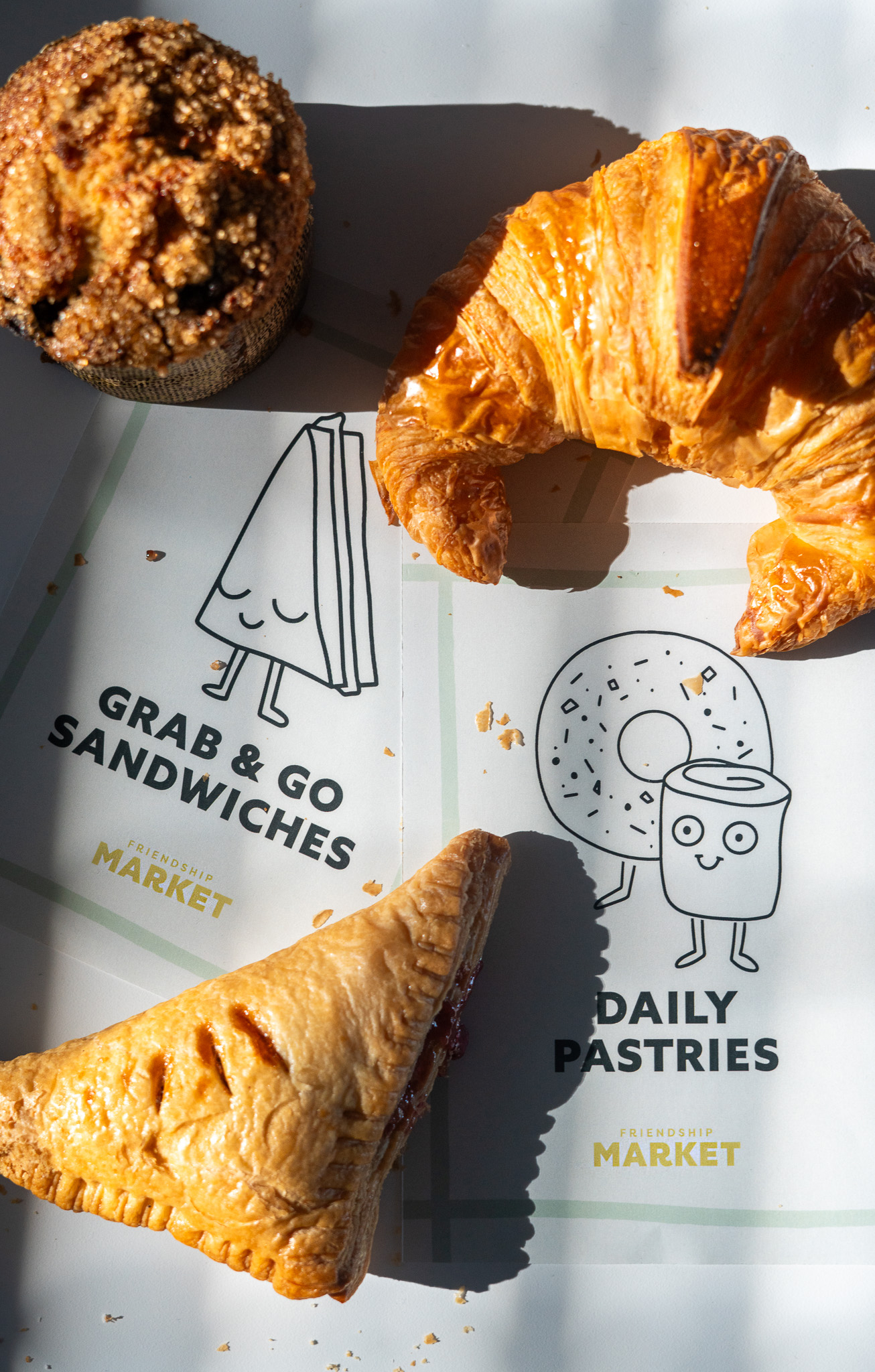 A croissant, a crumb-topped muffin, and a fruit turnover arranged on a surface with illustrated signs reading "Grab & Go Sandwiches" and "Daily Pastries," showcasing thoughtful brand strategy and design solutions.