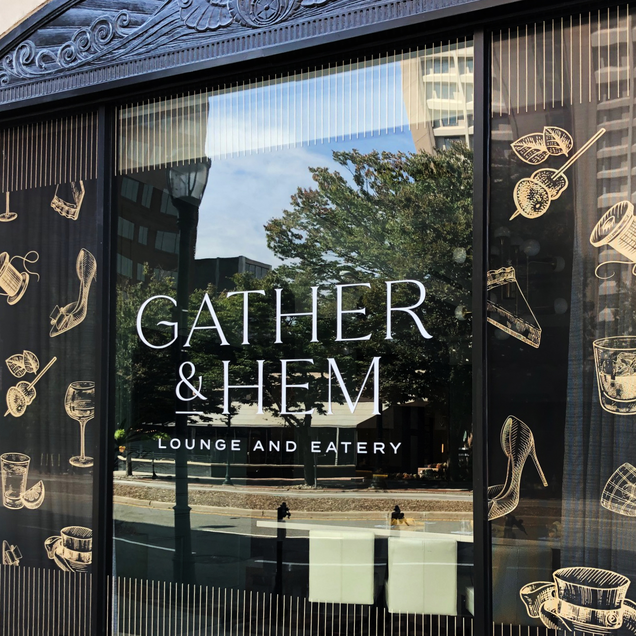 Storefront window displaying "Gather & Hem Lounge and Eatery," with illustrations of drinks and food—a showcase of thoughtful brand strategy reflected alongside trees and buildings in the glass.