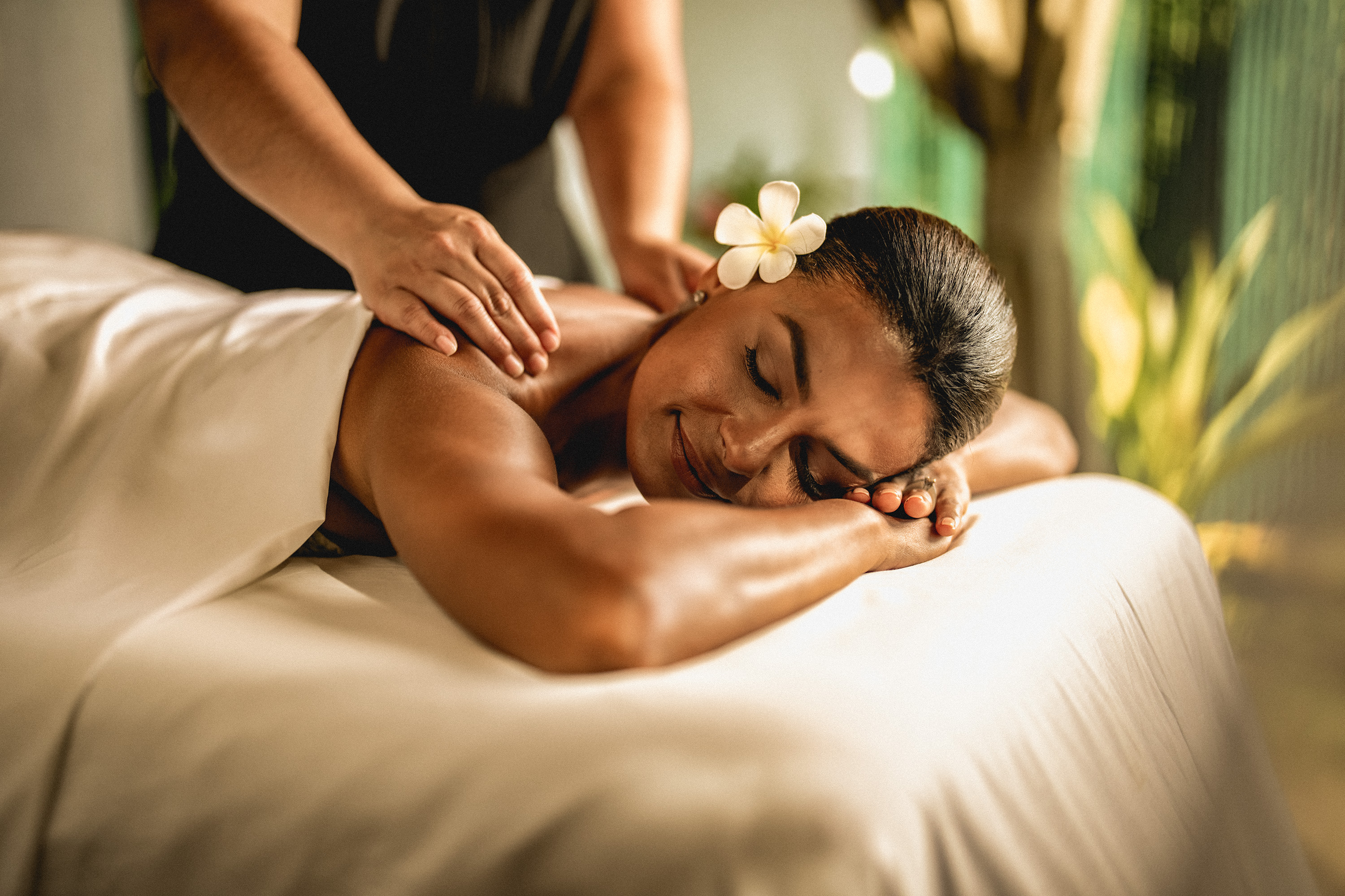 A woman with a flower in her hair lies on a massage table with her eyes closed, enjoying a shoulder massage in a spa setting that exemplifies soothing brand strategy and design solutions.