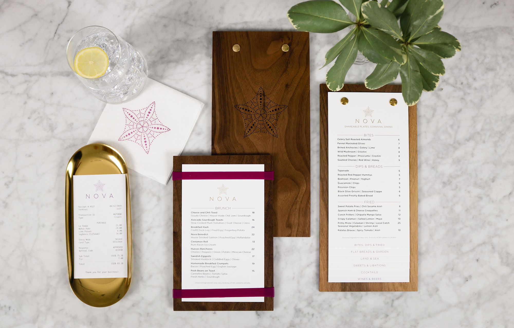 A marble table displaying restaurant menus on wooden boards, a small gold tray, a glass of water with lemon, a napkin, and a potted plant creates the perfect setting to reflect thoughtful brand strategy and design solutions.