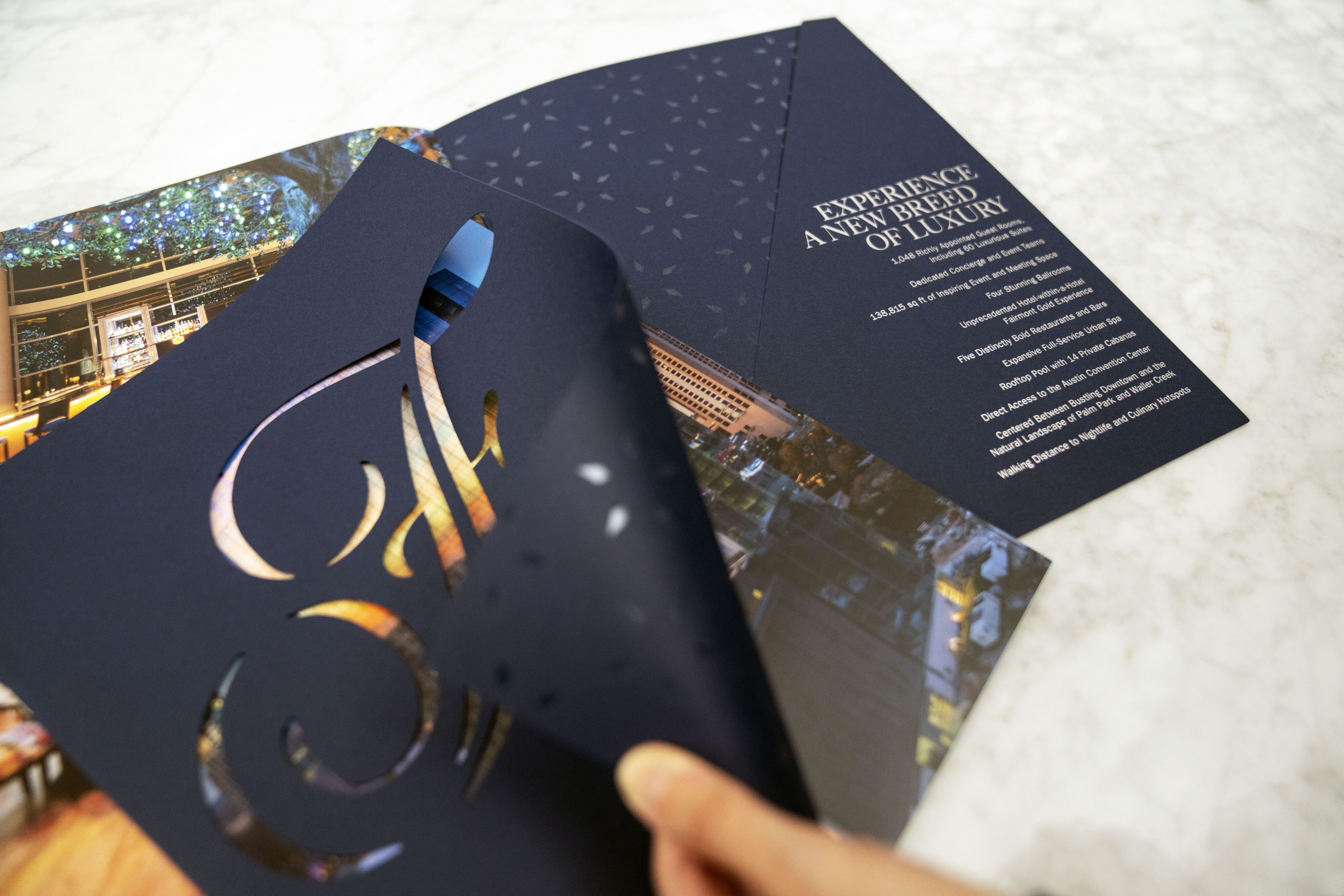 A hand lifts a dark blue brochure cover with a cut-out design, revealing how brand strategy and design solutions create an inner page of photos and the text “Experience a New Breed of Luxury” printed in white.