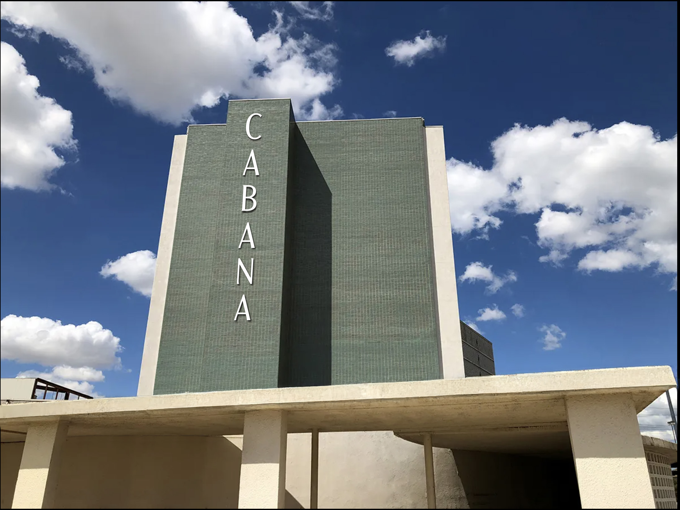 A building with a tall green wall displays the word "CABANA" in large vertical letters against a backdrop of a blue sky with scattered clouds.