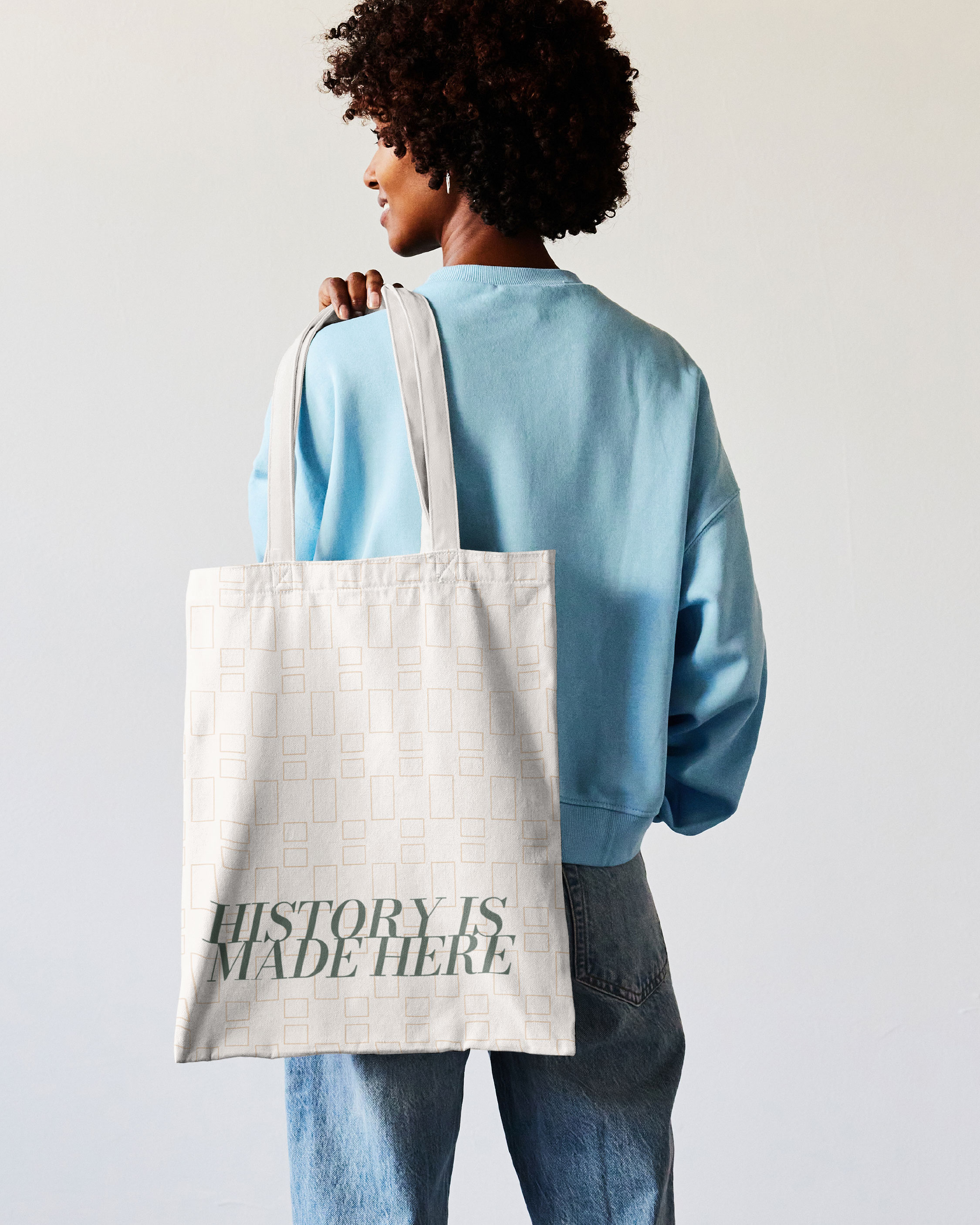 A person in a blue sweatshirt and jeans holds a white tote bag with a geometric pattern and the text "HISTORY IS MADE HERE.