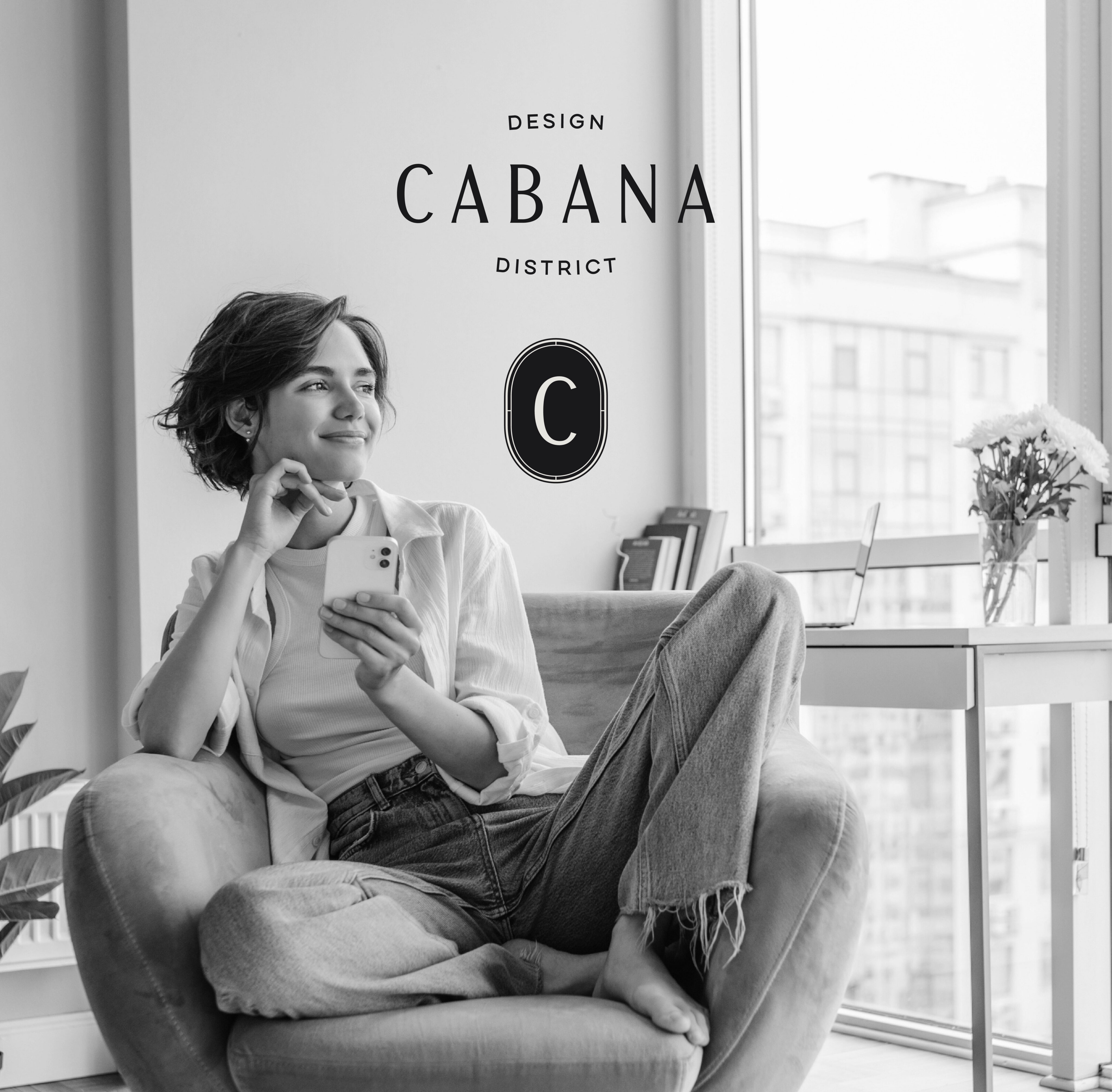 A person sits on a chair holding a smartphone and smiling, with a desk, flowers, and city buildings visible through large windows. Text reads: “Design Cabana District C.”.