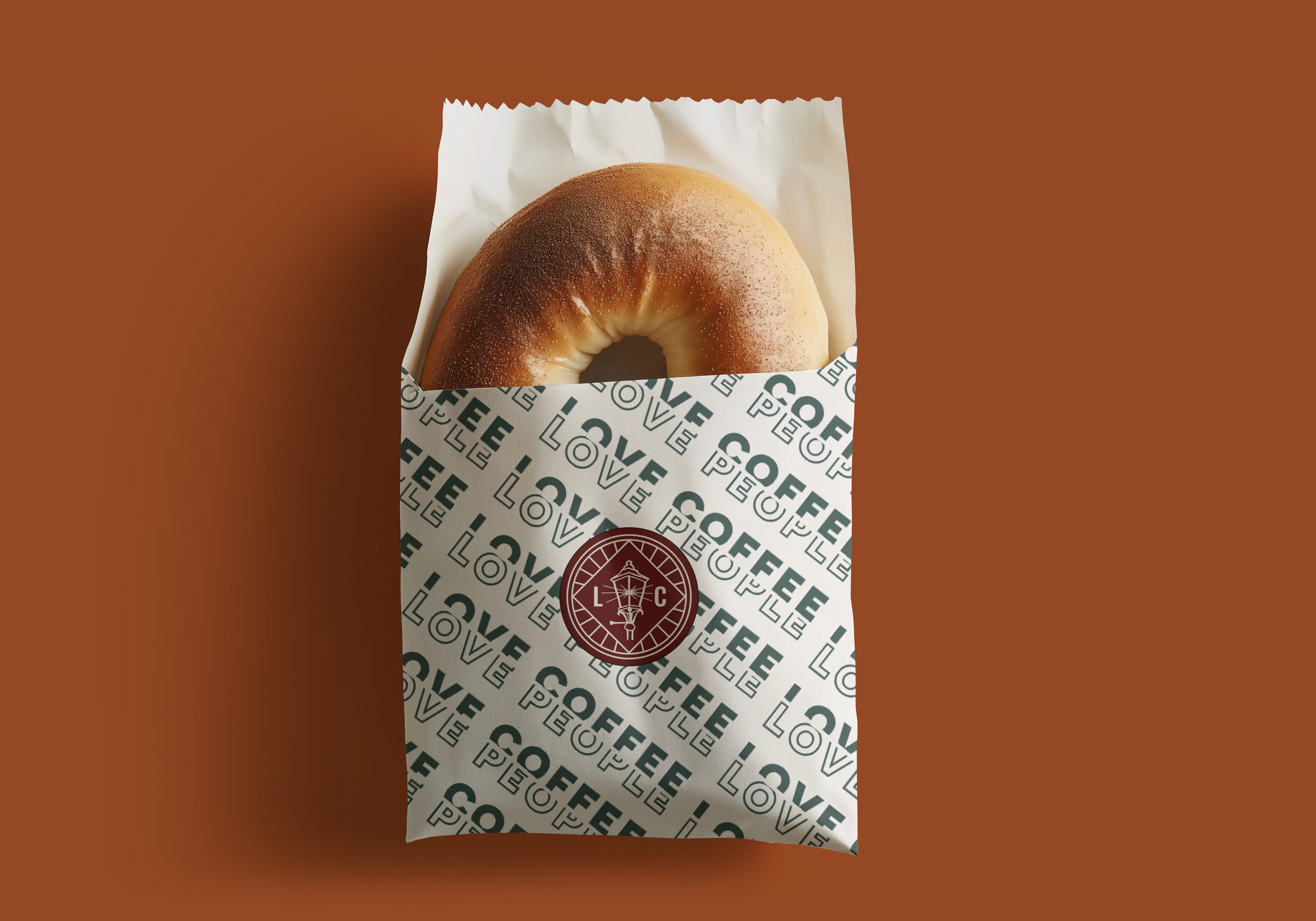 A plain bagel partially wrapped in branded paper with repeated text "LOVE COFFEE" and a circular logo, set against a solid brown background.