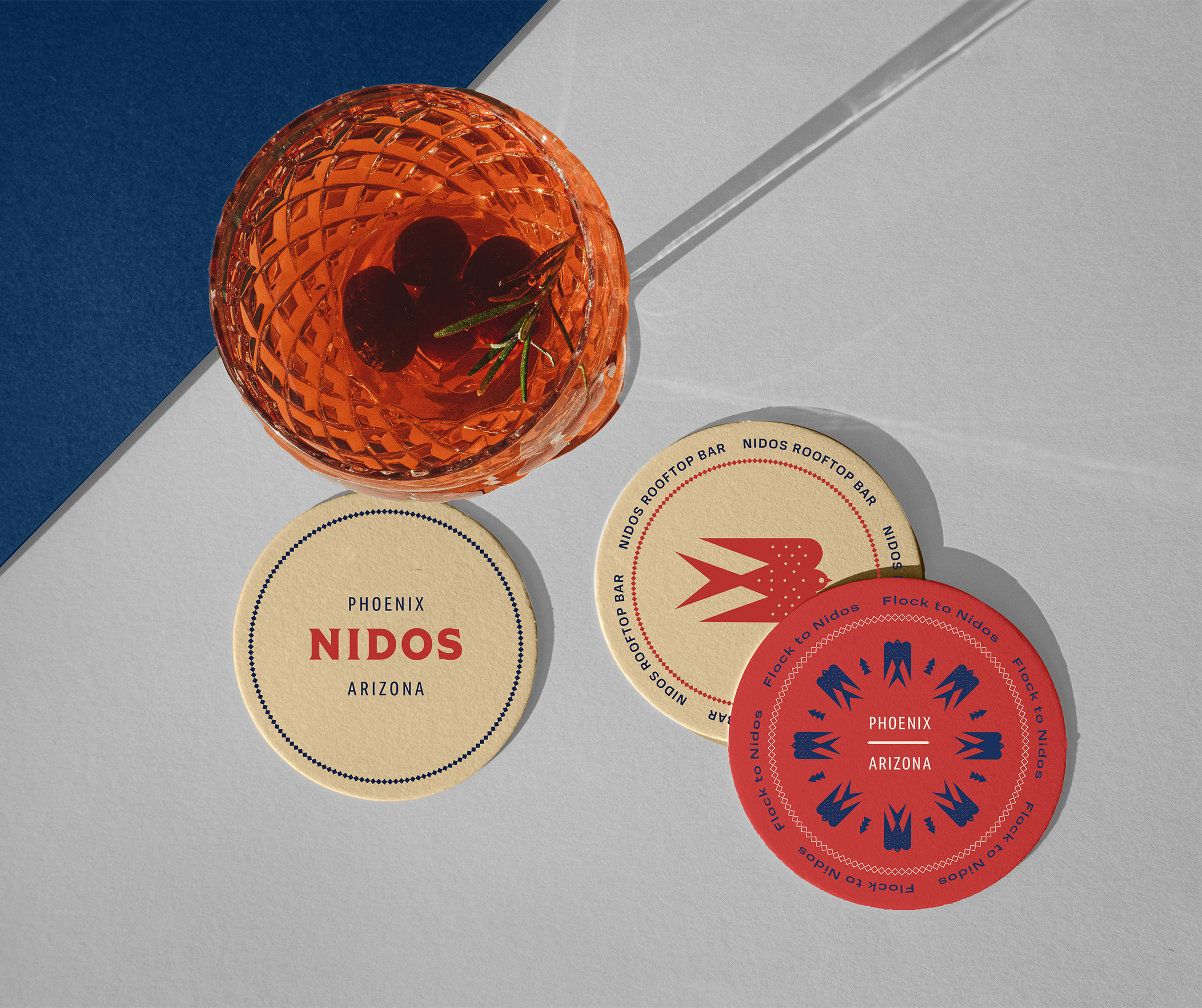 An orange cocktail with cherries sits on a white surface next to three round Phoenix Nidos Arizona branded coasters with graphic designs.