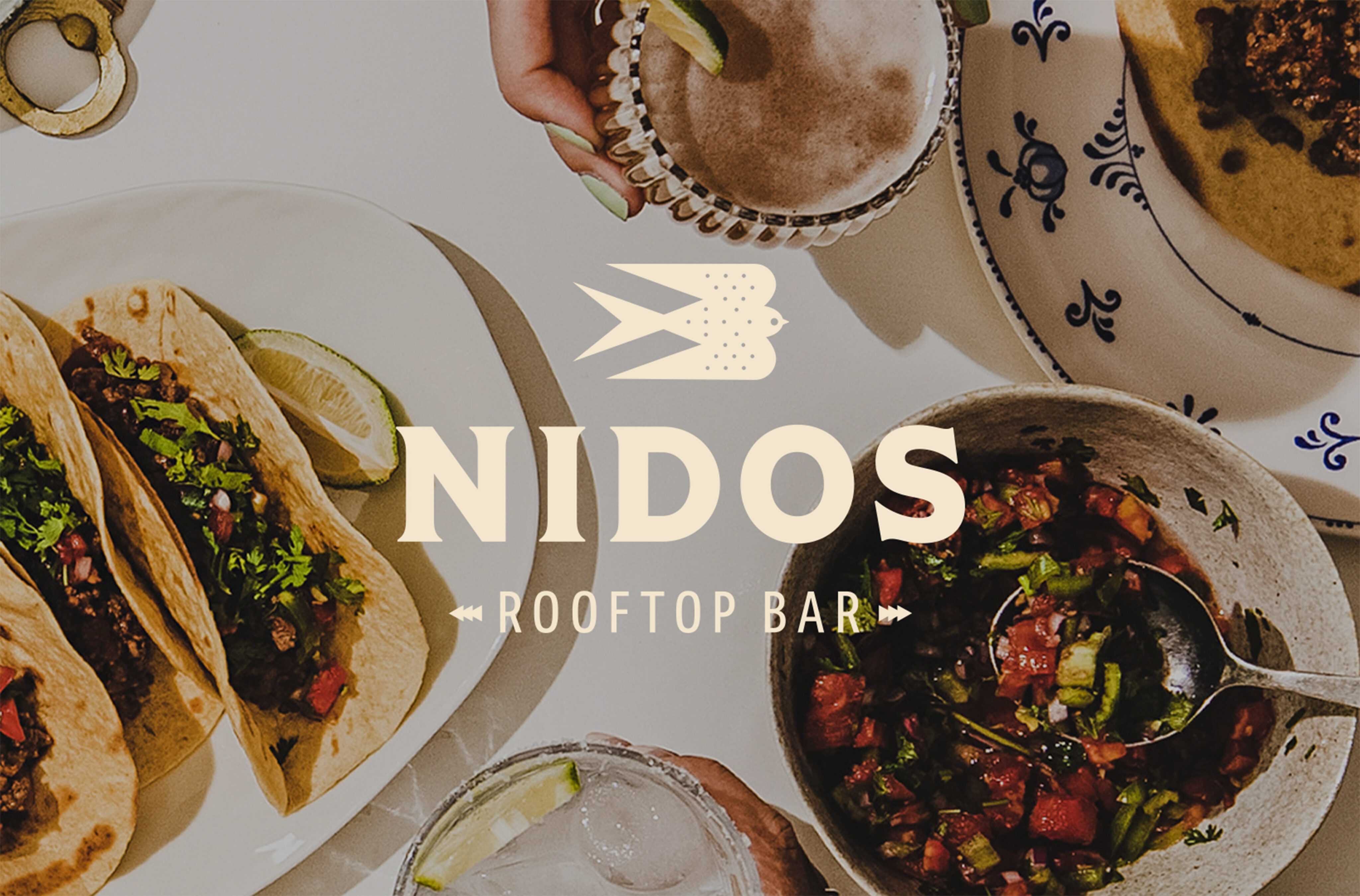 A table with tacos, salsa, drinks, and a hand holding a cocktail, overlaid with the text "NIDOS ROOFTOP BAR" and a bird logo.