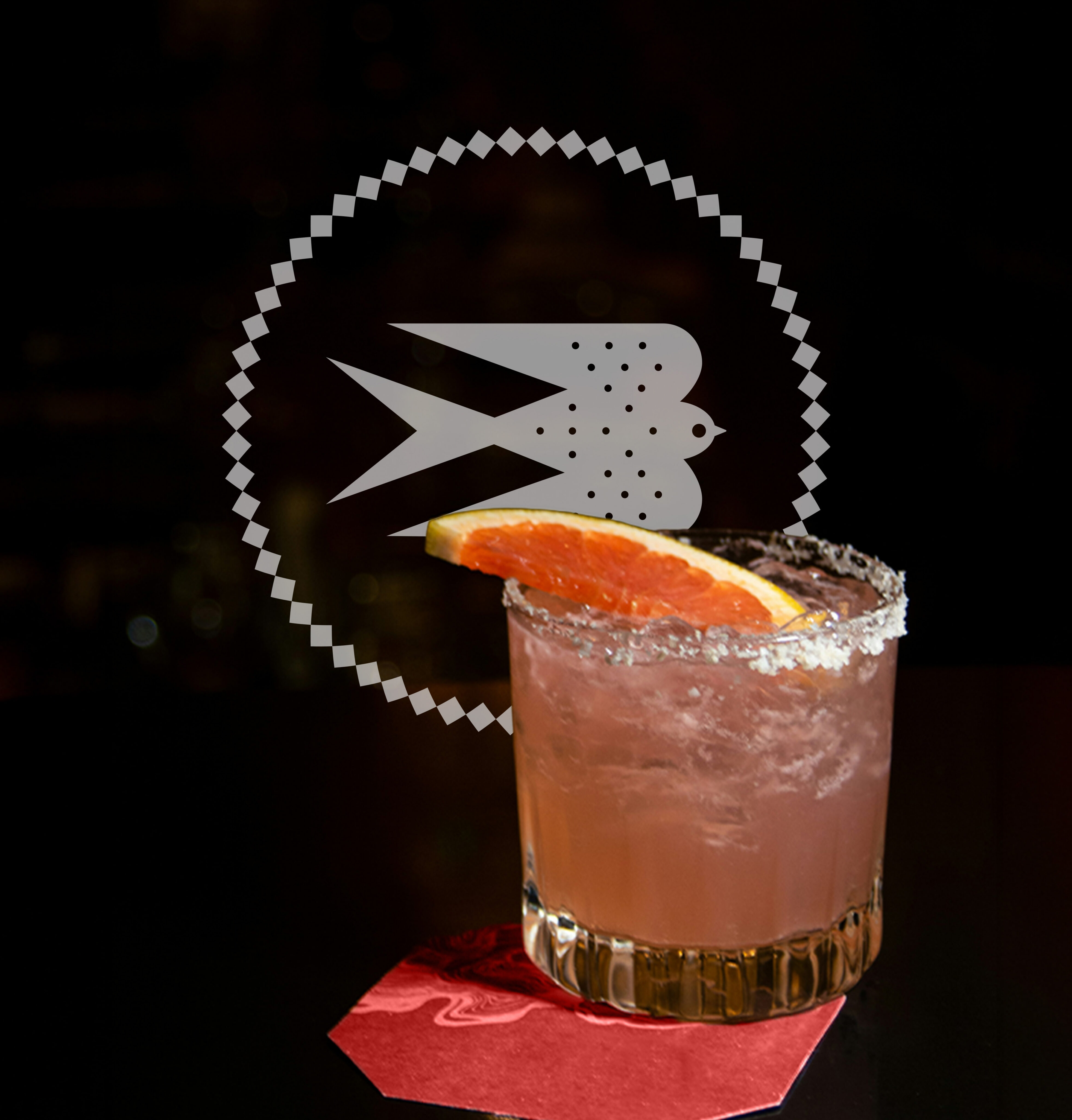 A cocktail with a salted rim and grapefruit wedge garnish sits on a red napkin; a bird logo is displayed in the background.