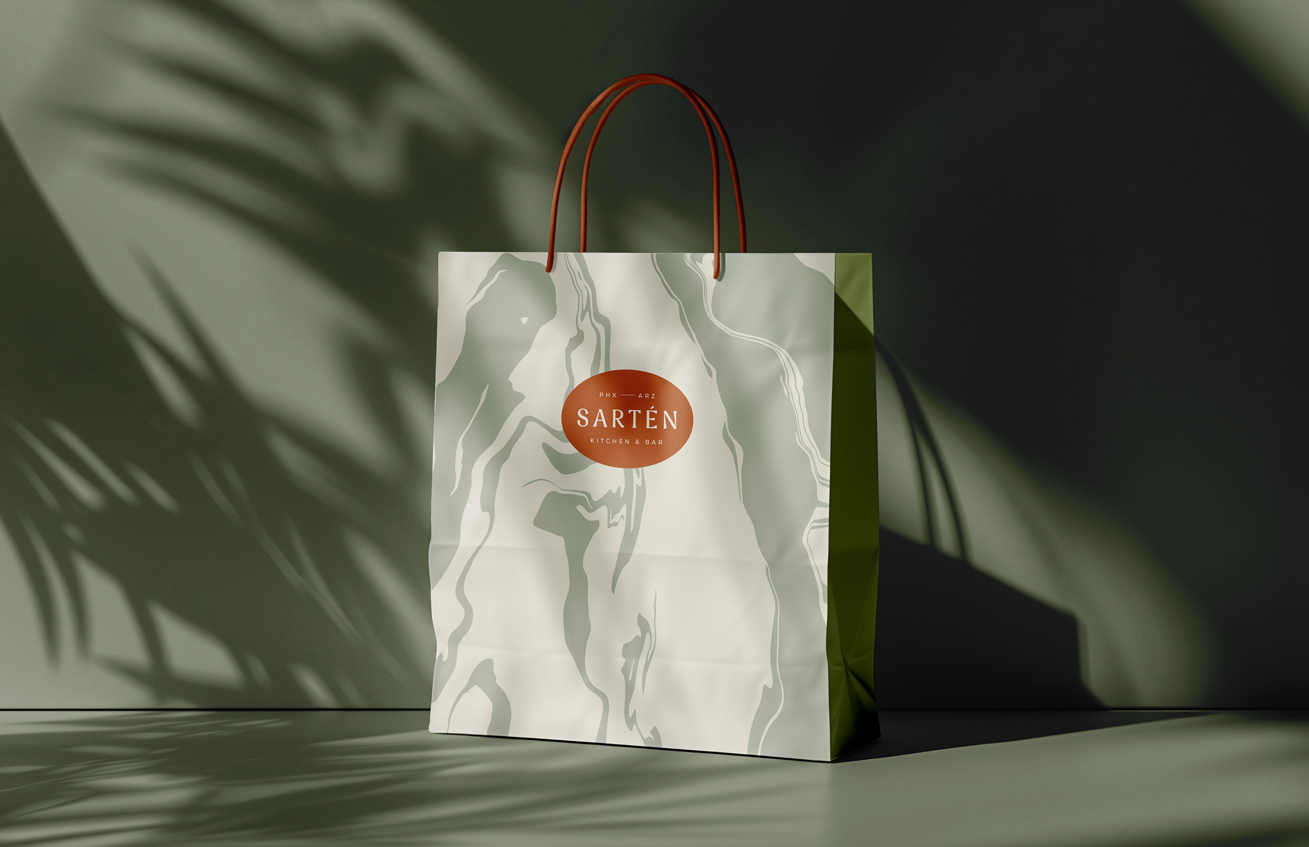 A paper shopping bag with red handles, featuring a marble pattern and an orange oval logo that reads "Sarten Kitchen & Home," is placed in a shadowy setting.