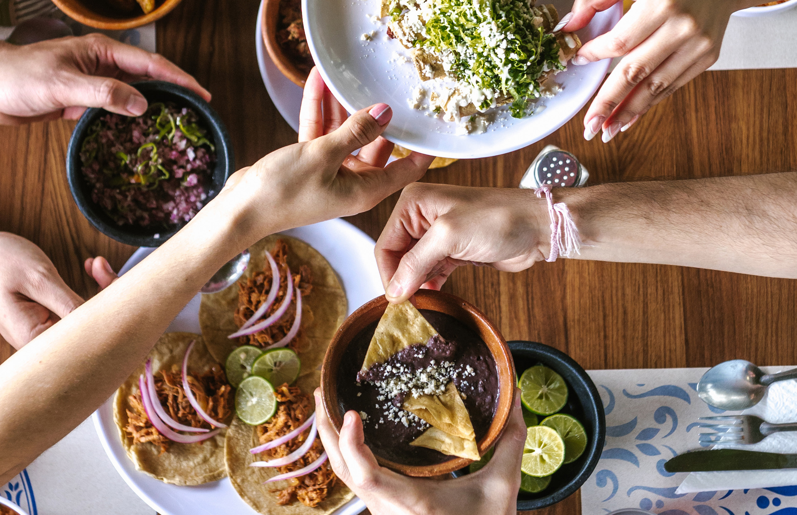 People sharing and reaching for various Mexican dishes, including tacos, bean dip with chips, and a bowl topped with cheese and herbs, on a wooden table.